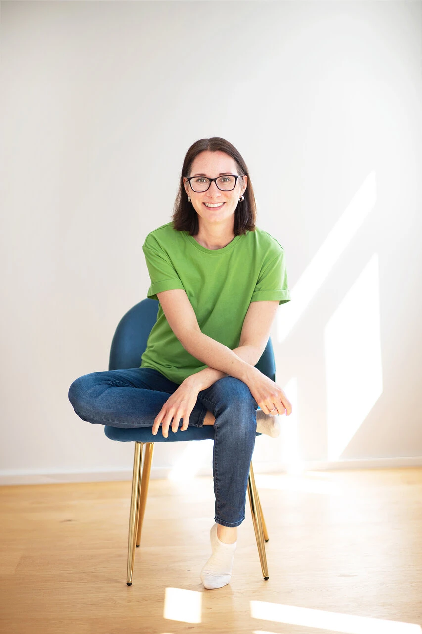 Karin Andre sitzt auf einem blauen Sessel, hat ein grünes Shirt und eine Bluejeans an. Das linke Bein ist lässig abgewinkelt auf der Sitzfläche und verschwindet unter dem rechten Bein, das auf dem Boden steht.